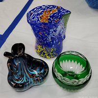 Photo of the three glass items: a speckled blue multicolor vase, a pear shaped dish with black and swirl blue/brown patterning, and an emerald green cut crystal candle holder.