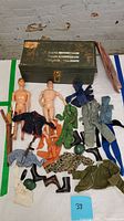 Two vintage GI Joe action figures with assorted clothing, boots, and toy weapons spread out next to a green metal storage box.