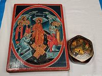 Both items side by side on a white cloth: the larger Greek religious painting on wood to the left and the smaller octagonal Russian lacquer vanity box to the right.