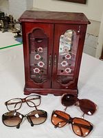 Full view of the dark wood vintage jewelry cabinet with floral glass doors and all four pairs of sunglasses arranged in front.