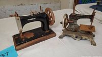 Two vintage miniature sewing machines side by side on display. One black marked Damascus, the other metal with floral decoration. Both aged with signs of rust and wear.