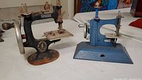 Photo of two vintage miniature sewing machines placed on a table: one black Singer branded and one blue machine.