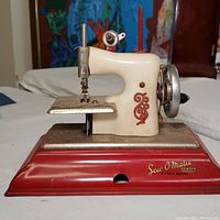 Front and side view of the vintage child's hand crank sewing machine showing red metal base, off-white body with red decoration, and hand crank on the side.
