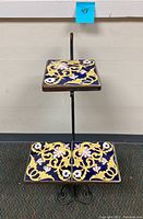 Front view of the plant stand showing two tile-covered tiers with intricate yellow and white floral patterns on blue background