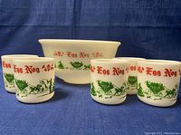 Punch bowl and five mugs with festive red 'Egg Nog' text and green holiday scenes on white milk glass, displayed against blue fabric background.