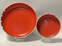 Front view of two round Mid-Century Modern ashtrays with orange enamel interiors on display stands, larger on left, smaller on right.