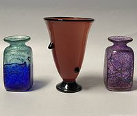 Three blown art glass vases displayed together: two rectangular bud vases in blue and aqua, and purple with blue speckles; one amber footed vase with black accents in the center.