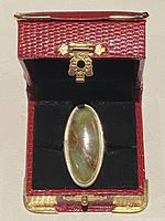 Top-down view of the silver ring with the oval stone sitting in a red jewelry box with gold-tone clasp and details.