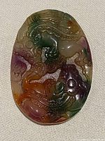 Front side of the oval jade pendant showing detailed carvings of mythical creatures and pronounced color variations including green, purple, orange, and white.