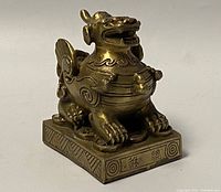 Angled side view of brass Foo Dog sculpture on square base featuring ruby jewel eyes and engraved Chinese characters on base edge.