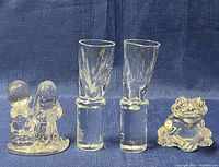 Two MCM-style shot glasses with thick bases, two Precious Moments figurines of children, and a Princess House gold-toned frog figurine on a blue fabric background.