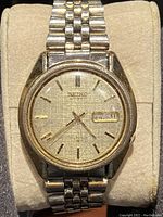 Front view of the Seiko automatic watch showing the textured gold-tone dial, day-date window displaying 'SAT 21', and stainless steel case and bracelet.