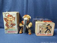 Front view showing the Tumbling Monkey toy in the box, blue Police Public Call Box tin lunch box, and Cracker Jack tin lunch box with clock and police children graphics.