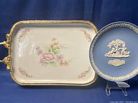 Rectangular footed serving tray with gold toned ornate metal frame and painted floral porcelain insert, alongside a blue Wedgwood Mother plate with white relief decoration.