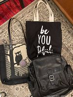 Overhead view of all items: black leather backpack, black tote bag with white text, beige tote bag with owl design, and small leather change purse