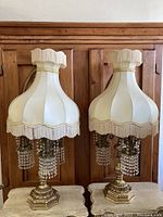 Full view of both vintage table lamps displayed side by side showing the total design with cream fabric bell-shaped shades trimmed in fringe and gold accents over gold-toned detailed bases with hanging crystal bead tassels.