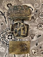 Three vintage metal belt buckles shown on patterned fabric background.