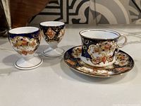 Demi tasse cup and saucer with two matching egg cups, showing ornate floral and geometric decorative pattern with gold trim.