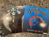 Front covers of The Steve Miller Band albums 'The Joker' (sealed) and 'Greatest Hits 1974-78' displayed on carpeted floor.