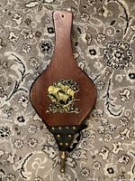 Wooden paddle-shaped fireplace bellows with black leather bottom and brass nozzle on patterned carpet background.