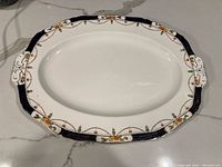 Top view of oval vintage Alfred Meakin platter showing floral and scroll decorative border and integrated handles. White ceramic base with dark blue, orange, and gold accent details.