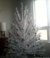 Assembled 7' aluminum Christmas tree displayed with star topper and reflective ornaments in front of window. Silver tinsel branches on metal pole sections; plastic tripod stand visible.