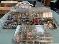Overview of three plastic compartment cases showing a variety of pins, earrings, and chains organized in individual compartments