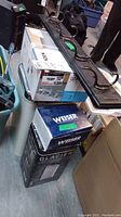 Lot items positioned together showing the unopened boxes for Moen faucet, Weiser lock, Glacier light fixture, black metal coat hanger with five hooks, and plastic garbage can.