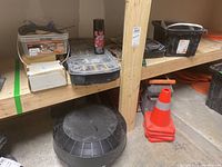Photo shows wooden shelving with small plastic containers holding screws, a can of spray, a black plastic roof exhaust vent on the floor, and stacked orange traffic pylons beneath shelf.