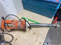 Photo showing the orange and silver hand mixer laid flat on a wooden surface, highlighting the mixing shaft and handle.