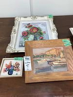 Overview of three framed artworks on a table: floral oil painting, harbor scene painting, and small floral print.