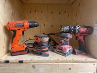 Four power tools on wooden shelf: Milwaukee M18 cordless drill, Milwaukee corded palm sander, Black & Decker 20V cordless drill, Black & Decker corded palm sander.