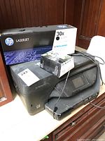 HP OfficeJet Pro 6978 printer with print cartridge and HP branded 30X LaserJet toner cartridge box on top, shown with cords and surrounding furniture.