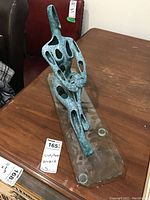 Abstract turquoise-green ceramic sculpture with multiple cutout holes on a rectangular glass plinth, shown on a wooden table.