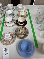 Overview of nine teacup and saucer sets on table; mix of floral patterns and colors, some with gold trim; includes light blue Pyrex set