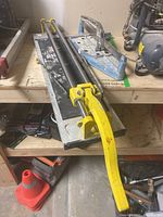 Large manual tile cutter with yellow handle, metal rails, black base, visible wear, placed on wooden shelf
