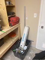 Photo showing black plastic container filled with boxed Moen bathroom fixtures and assorted tools, positioned against wall near wooden shelving.