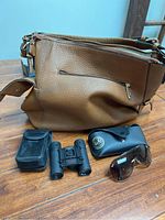 Photo of brown faux leather purse with zipper and strap, compact binoculars, black carrying case and sunglasses on table.