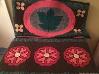 Two vintage hand hooked rugs stacked, visible are a rug with a green maple leaf in a pink oval and floral corners, and a rug with three pink flowers