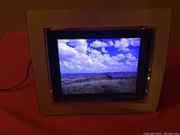 Front view of digital photo frame displaying an outdoor landscape image with blue sky and clouds.