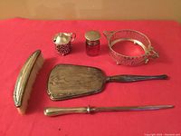 Full lot arranged on red cloth showing all six pieces: a hand brush, vanity mirror, small creamer, glass bottle with lid, circular pierced dish, and letter opener.