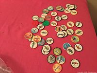 Overview of approximately 45 vintage Jello collector coins scattered on a red surface showing colorful vintage automotive illustrations.