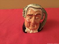 Front view of the Royal Doulton Toby mug showing the detailed hand-painted face and feather pen handle on a red background.