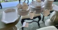 Wide angle photo showing full lot on table, displaying stacked plates, cups, bowls, serving platter, creamer, and sugar bowl all in white fine china with embossed floral border.