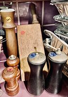 Wooden salt and pepper grinders, wooden and black salt and pepper shakers, and wooden cutting board arranged near metal trays.