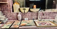 View of large ceramic bowls with palm tree design, large jug, white vase, and four framed kitchen-themed pictures on countertop.