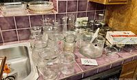 Collection of various clear glass kitchenware items including Kerr jars, bowls, pie pans, vases, and a salad bowl with utensils arranged on a countertop.