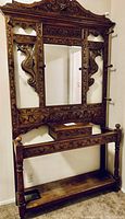 Front view of ornate carved wood hall tree with central mirror, coat hooks, hinged storage box and lower shelf.