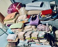Full overview of stacked assorted linens including towels, blankets, comforters, fabric pieces