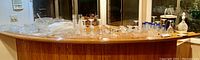 Wide view of the assorted glassware displayed on a wooden counter, including glasses, decanters, ice bucket, coasters, platters, and a punch bowl set.